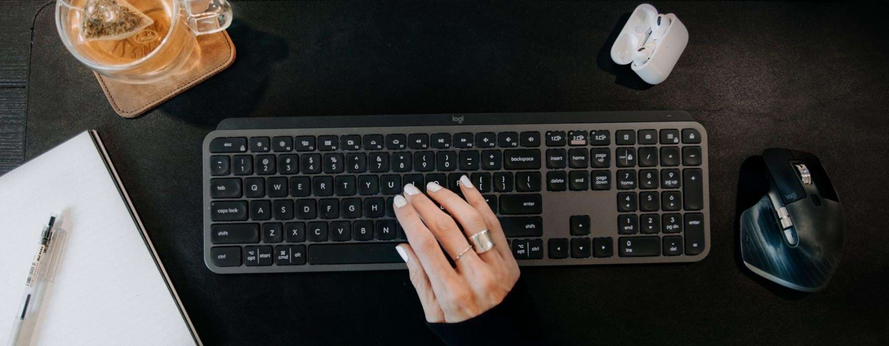 woman's hand on a keyboard surrounded by office items and a cup of tea on a coaster